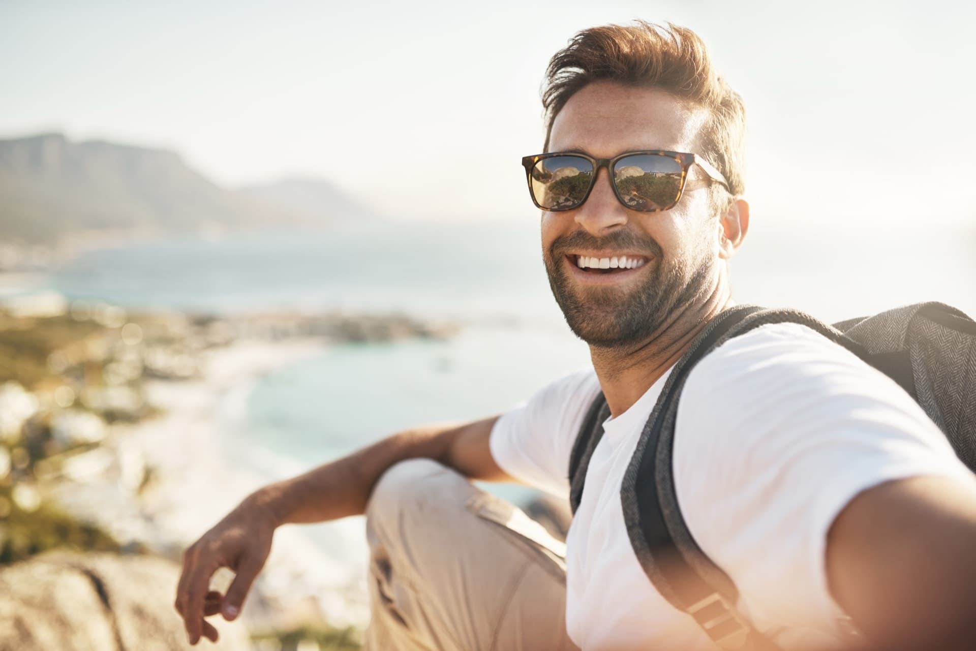 A smiling man in sunglasses resting on a hillside above a sunlit coastline
